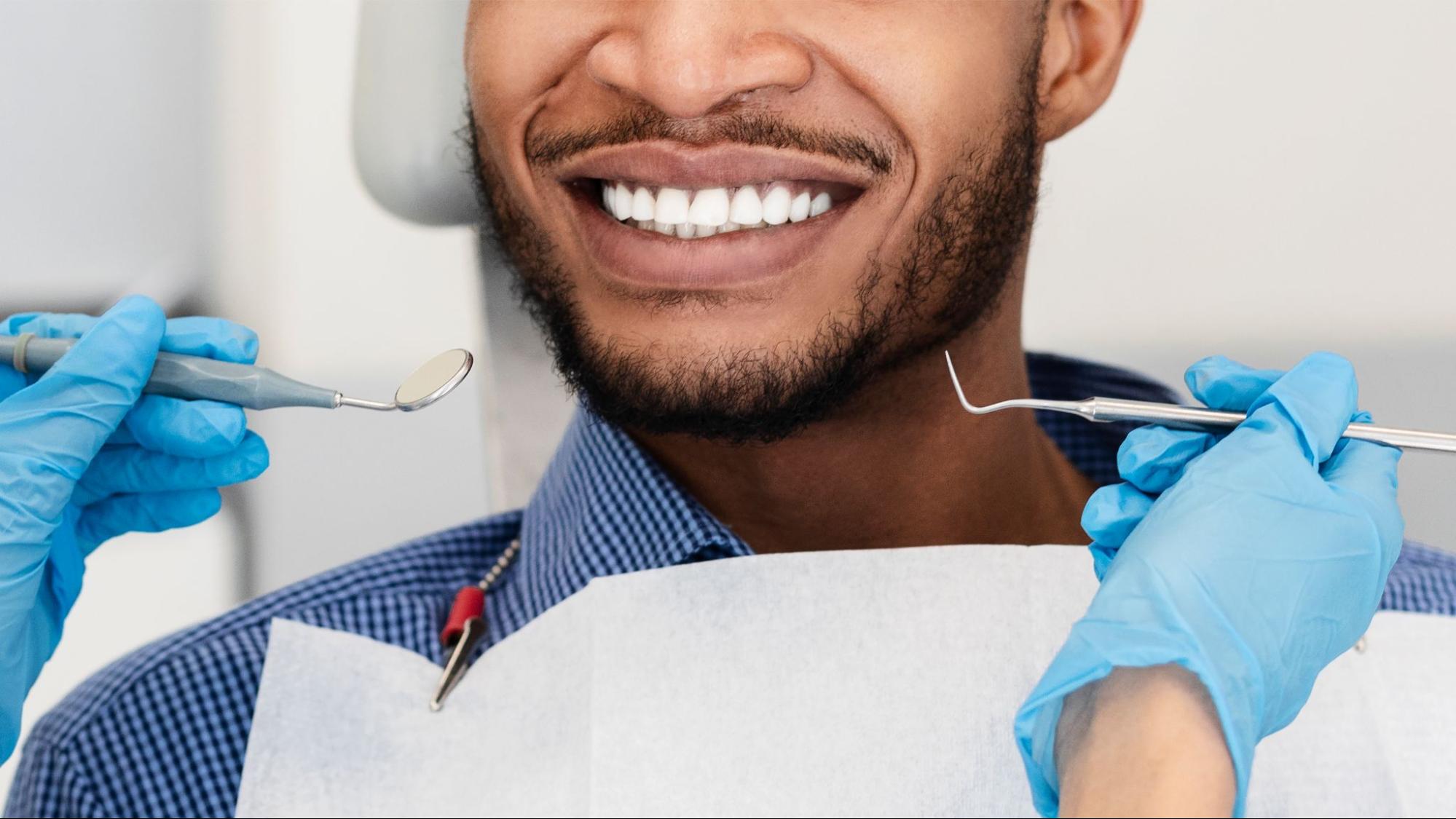 Person with white teeth smiling next to a dentist with gloves holding tools.