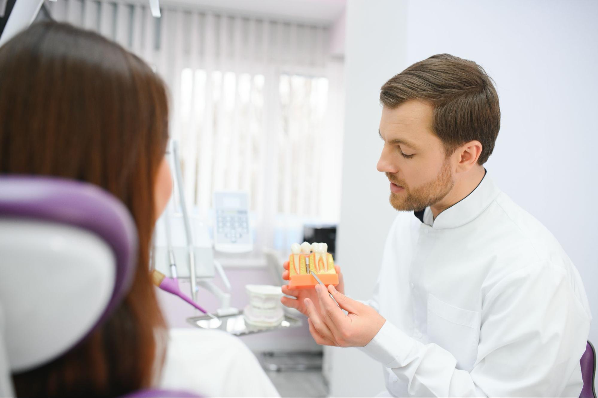 A male dentist demonstrates the steps for a dental implant procedure using a plastic tooth implant model.