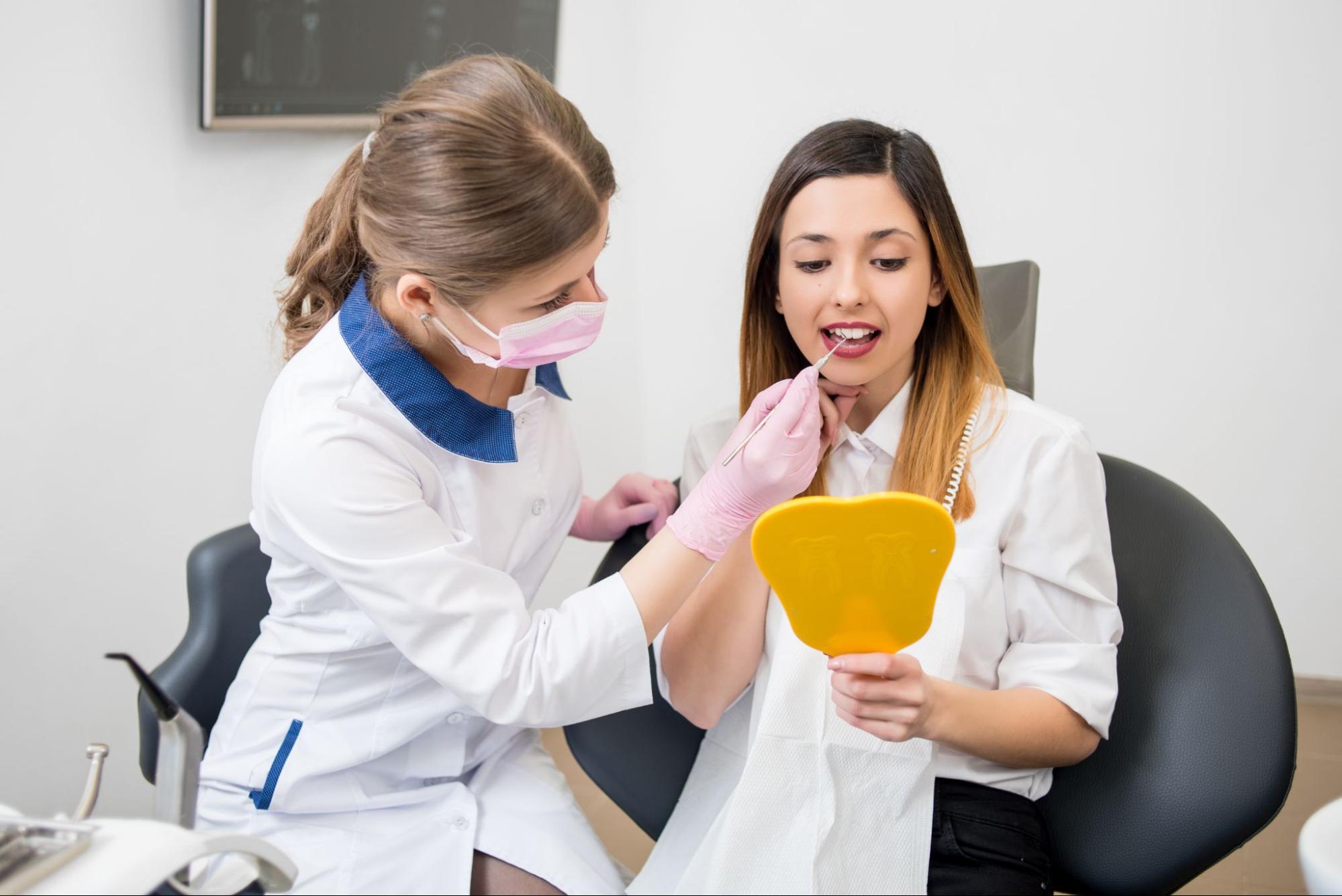 A female dentist examines a woman's teeth with a dental probe while the patient looks at herself in a mirror after a plaque cleaning.