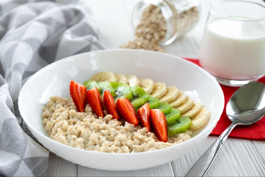 A bowl of soft oatmeal topped with sliced strawberries, bananas, and kiwis, served with a glass of milk.