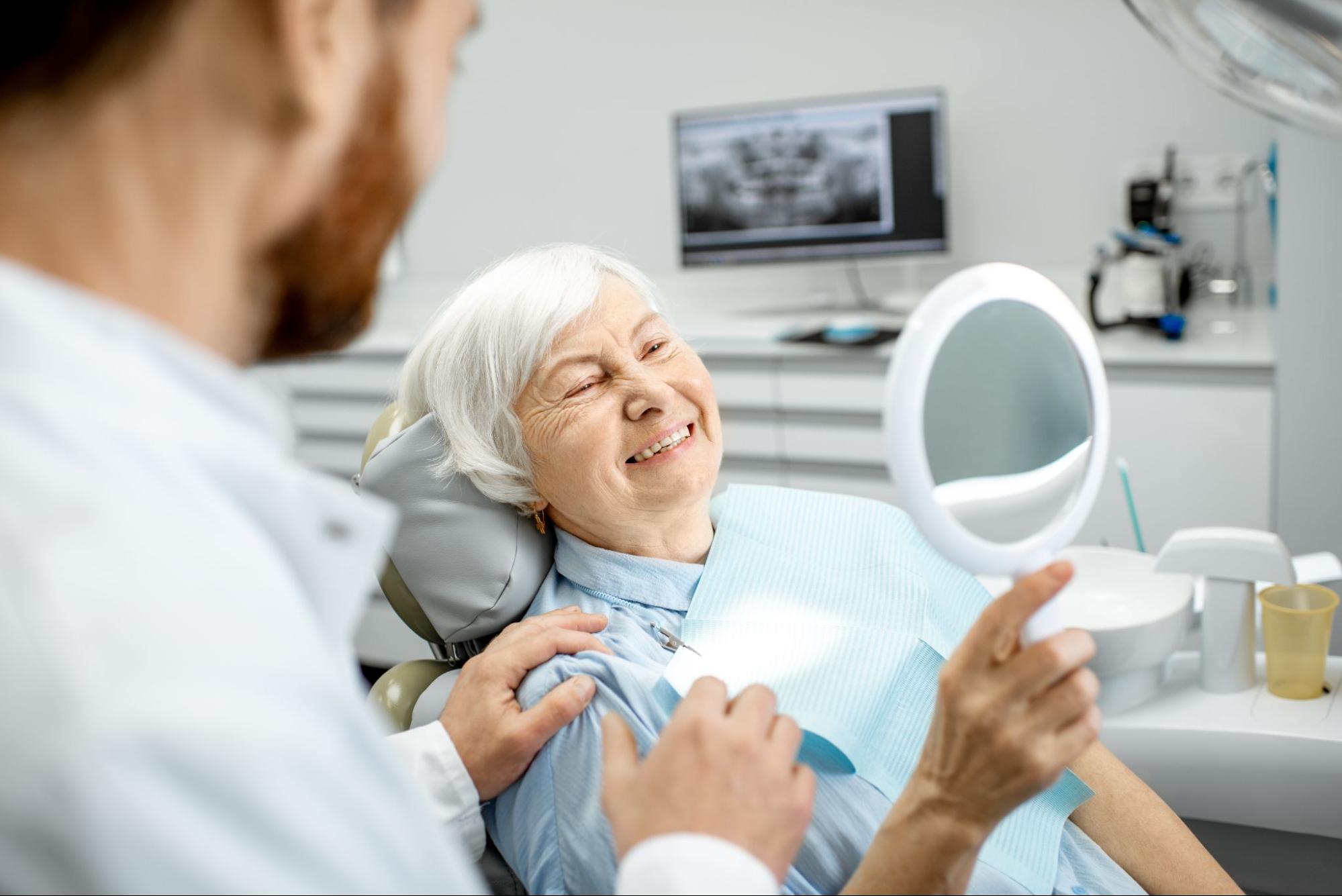 An older woman admires her new dentures in the mirror as the dentist observes.