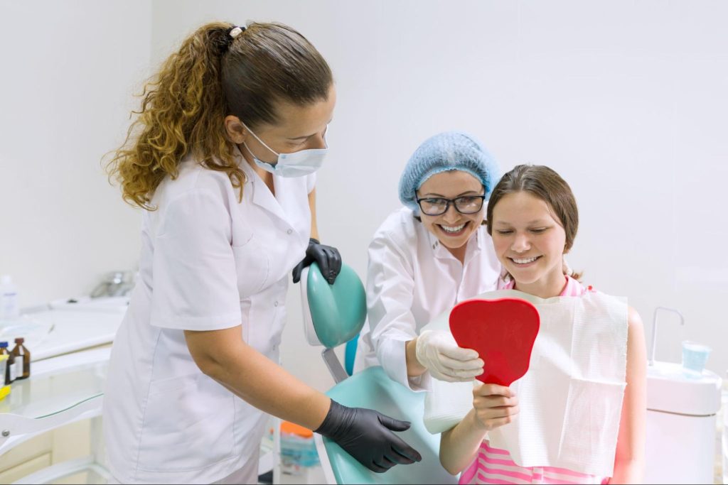A happy teenage girl checks her teeth in a handheld mirror after a dental procedure.